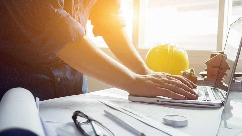 An engineer works on a laptop computer with his yellow hard hat sitting next to it.